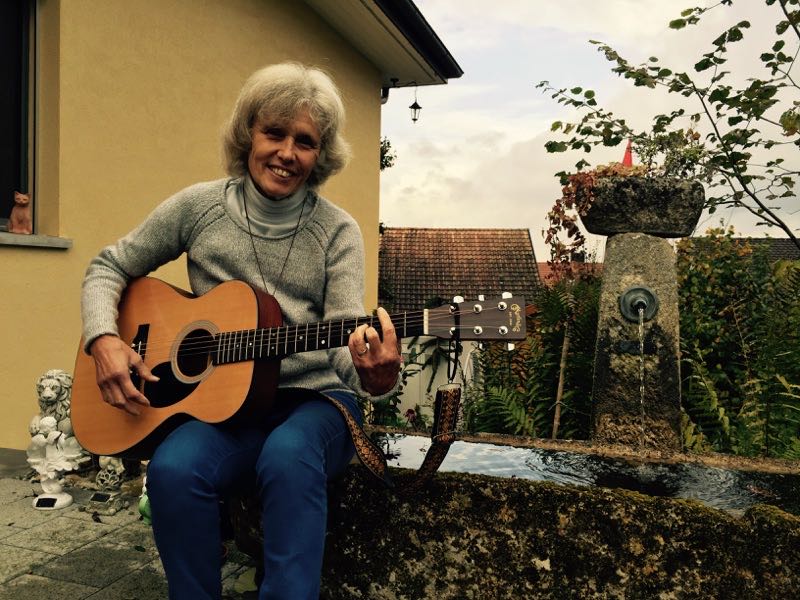 Christina Ueberwasser Koch mit Gitarre am Brunnen vor dem Haus